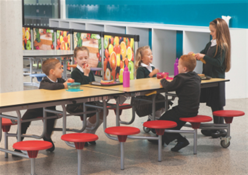 A group of children are sat on red, plastic stools that have been attached to a maple school dining table. The table is a 12 seater.